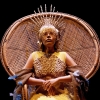 Woman wearing a gold dress and crown sits in a straw thrown chair 